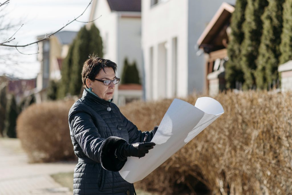 pexels-photo-7937208-7937208 Woman examining a blueprint in a sunny residential area, considering properties.