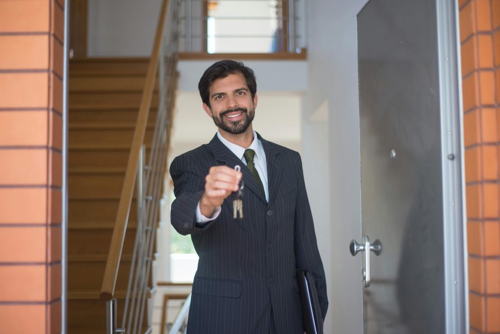 pexels-photo-8815910-8815910 Smiling realtor in a black suit handing keys through an open door inside a modern house.